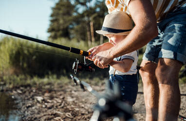 A midsection of father with small toddler boy fishing by a lake, holding a rod. - HPIF20187