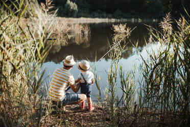 Eine Rückansicht eines reifen Vaters mit einem kleinen Sohn im Kleinkindalter, der an einem Fluss oder See angelt. - HPIF20196