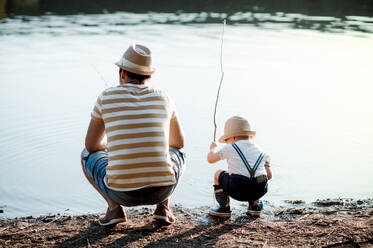 Eine Rückansicht eines reifen Vaters mit einem kleinen Sohn im Kleinkindalter, der an einem Fluss oder See angelt. - HPIF20204