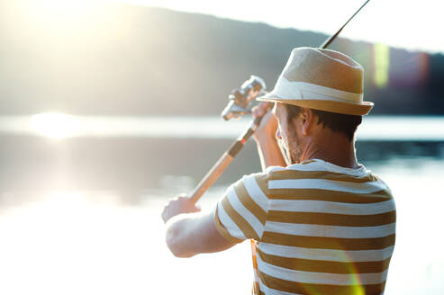 Ein reifer Mann mit Hut, der bei Sonnenuntergang an einem See angelt und eine Angelrute in der Hand hält. copy space. - HPIF20205