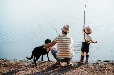 Eine Rückansicht eines erwachsenen Vaters mit einem kleinen Sohn und einem Hund, der an einem Fluss oder See angelt. - HPIF20206
