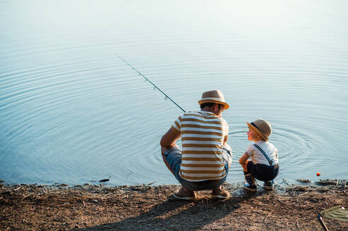 Eine Rückansicht eines reifen Vaters mit einem kleinen Sohn im Kleinkindalter, der an einem Fluss oder See angelt. - HPIF20207