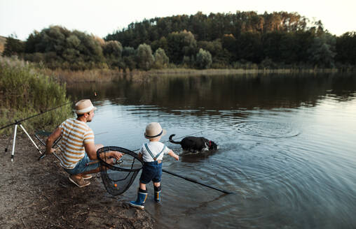 Ein erwachsener Vater mit einem kleinen Sohn im Kleinkindalter und einem Hund beim Angeln an einem Fluss oder einem See. - HPIF20217