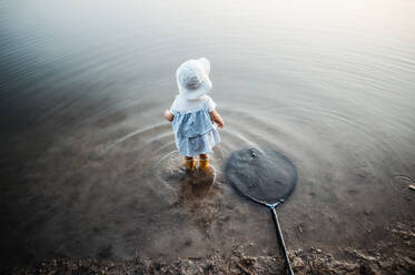 Blick von hinten und von oben auf ein kleines Kleinkind, das in einem See im Wasser steht, mit einem Fischernetz neben sich. - HPIF20221