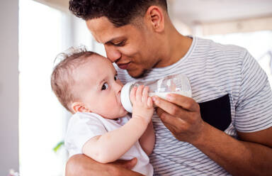 A portrait of cheerful father bottle feeding a small toddler son indoors at home. - HPIF20228