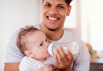 A portrait of cheerful father bottle feeding a small toddler son indoors at home. - HPIF20231