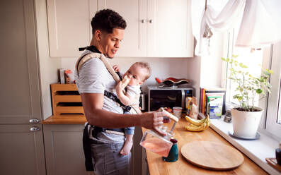 A father with small toddler son in carrier in kitchen indoors at home, pouring tea. - HPIF20232