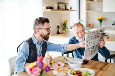 Ein erwachsener Hipster-Sohn und ein älterer Vater sitzen zu Hause am Tisch, essen ein leichtes Mittagessen und lesen Zeitung. - HPIF20306