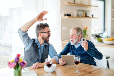 Adult hipster son and senior father with wine and tablet sitting at the table indoors at home. - HPIF20311