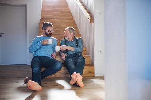 Ein erwachsener Hipster-Sohn und ein älterer Vater sitzen mit einem Kaffee am Fuße einer Treppe in einem Haus und unterhalten sich. - HPIF20356