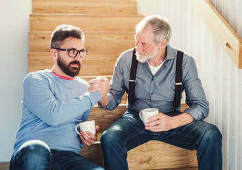 Ein erwachsener Hipster-Sohn und ein älterer Vater sitzen mit einem Kaffee am Fuße einer Treppe in einem Haus und unterhalten sich. - HPIF20357