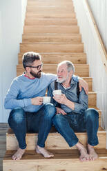 Ein erwachsener Hipster-Sohn und ein älterer Vater sitzen mit einem Kaffee am Fuße einer Treppe in einem Haus und unterhalten sich. - HPIF20359