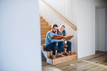 Ein erwachsener Hipster-Sohn und ein älterer Vater sitzen zu Hause auf einer Treppe und schauen sich Fotos an. - HPIF20363