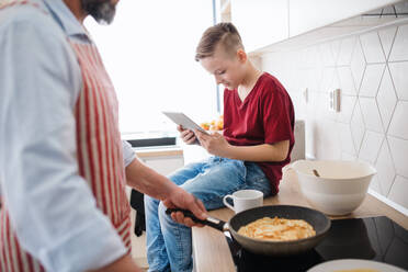 A mature father and small son with tablet indoors in kitchen, making pancakes. - HPIF20390
