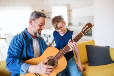 Ein reifer Vater mit seinem kleinen Sohn sitzt drinnen auf dem Sofa und spielt Gitarre. - HPIF20398