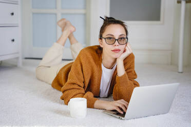 Freelancer with coffee cup and laptop lying on carpet at home - NLAF00037