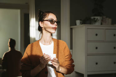 Nachdenkliche Frau mit Sonnenlicht im Gesicht, die zu Hause eine Kaffeetasse hält - NLAF00039