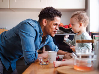 A father and a small toddler son eating fruit and yoghurt in kitchen indoors at home. - HPIF20416