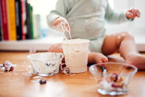 Ein Mittelteil eines kleinen, unordentlichen Kleinkindes, das zu Hause auf dem Küchentisch sitzt und Obst und Joghurt isst. - HPIF20417