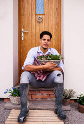 Ein Porträt des jungen fröhlichen Mannes Gärtner sitzt vor der Tür zu Hause, hält Korb mit Blumen. - HPIF20446