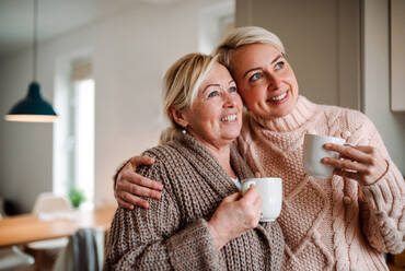 A happy senior woman and adult daughter with coffee at home, looking out of window. - HPIF20489