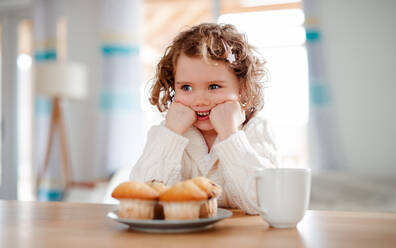 Ein Porträt eines glücklichen kleinen Mädchens mit Muffins, das zu Hause am Tisch sitzt. - HPIF20495