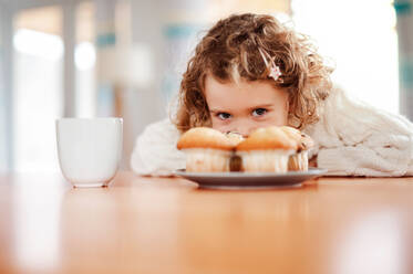 Ein Porträt eines glücklichen kleinen Mädchens mit Muffins, das zu Hause am Tisch sitzt. - HPIF20496