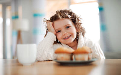 Ein Porträt eines glücklichen kleinen Mädchens, das zu Hause am Tisch sitzt und Muffins isst. - HPIF20498