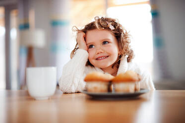 Ein Porträt eines glücklichen kleinen Mädchens mit Muffins, das zu Hause am Tisch sitzt. - HPIF20499