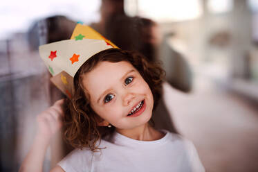A portrait of small girl with a paper crown at home, looking at camera. Shot through glass. - HPIF20513