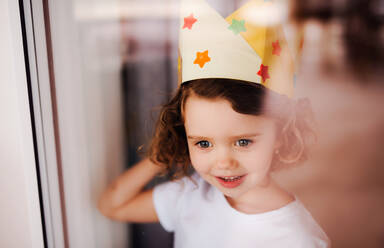 A portrait of small girl with paper crown on head at home, looking out of a window. Shot through glass. - HPIF20514