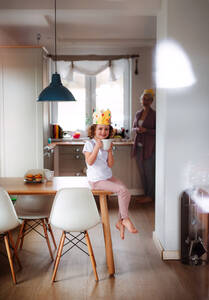 Seitenansicht eines kleinen Mädchens und einer Mutter mit Papierkrone zu Hause, beim Trinken. - HPIF20520