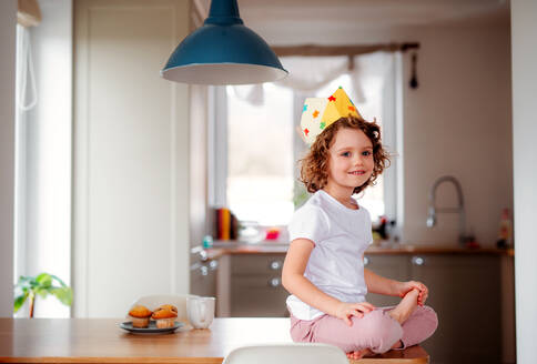 Ein Porträt eines kleinen Mädchens mit einer Papierkrone zu Hause, das in die Kamera schaut. - HPIF20521