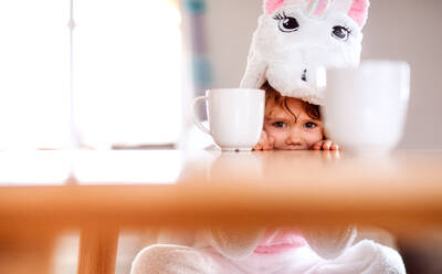 Ein Porträt eines glücklichen kleinen Mädchens mit Einhornmaske, das zu Hause am Tisch sitzt. - HPIF20529