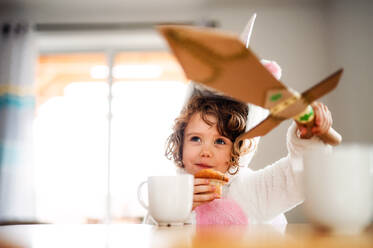 Ein Porträt eines glücklichen kleinen Mädchens mit einem Papierschwert, das zu Hause am Tisch sitzt und Muffins isst. - HPIF20535