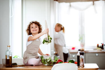 Porträt eines kleinen Mädchens mit Großmutter in einer Küche zu Hause, die einen Gemüsesalat zubereitet. - HPIF20549