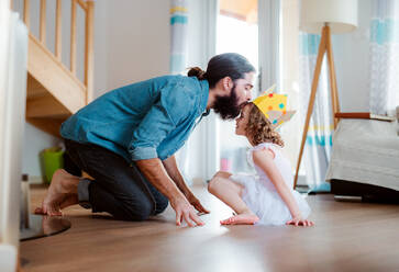 Seitenansicht eines kleinen Mädchens mit einer Prinzessinnenkrone und einem jungen Vater zu Hause, die sich beim Spielen küssen. - HPIF20593