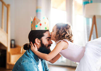Seitenansicht eines kleinen Mädchens mit einer Prinzessinnenkrone und einem jungen Vater zu Hause, die sich beim Spielen küssen. - HPIF20594