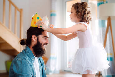 A side view of small girl putting a paper crown on father's head at home when playing. - HPIF20596
