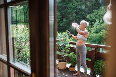 Eine ältere Frau mit Sport-BH auf einer Terrasse im Sommer, Wasser trinkend, Aufnahme durch Glas. - HPIF20620