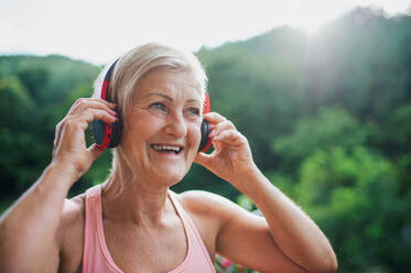 Eine ältere Frau mit Kopfhörern steht im Sommer draußen auf einer Terrasse und hört Musik. - HPIF20622