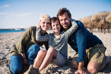 A young family with small boy sitting outdoors on beach, looking at camera. - HPIF20649