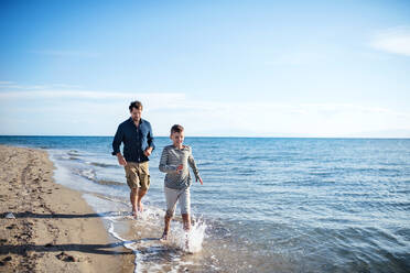 Father with small son on a walk outdoors on beach, running barefoot in water. - HPIF20650