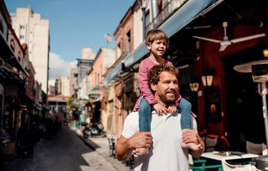 Vater mit kleiner Tochter steht im Freien auf der Straße in einer mediterranen Stadt und nimmt sie Huckepack. - HPIF20701