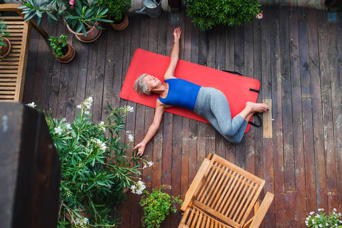 Draufsicht auf eine ältere Frau auf einer Terrasse im Sommer, die auf einer Matte Übungen macht. - HPIF20743
