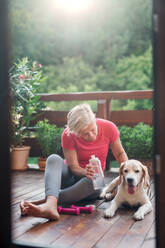 Eine ältere Frau mit Hund auf einer Terrasse im Sommer, die sich nach dem Sport ausruht. - HPIF20757