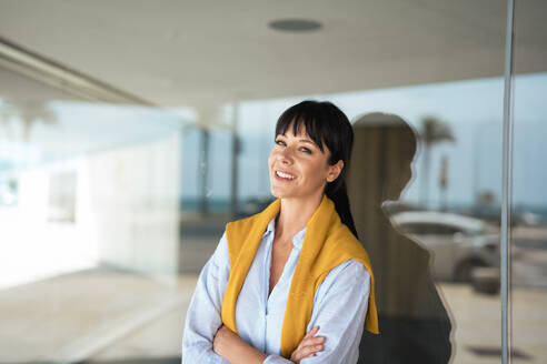 Smiling mature businesswoman with arms crossed near glass wall - JOSEF19615