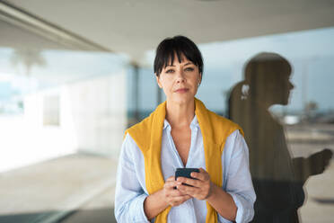 Confident businesswoman holding mobile phone in front of glass wall - JOSEF19630
