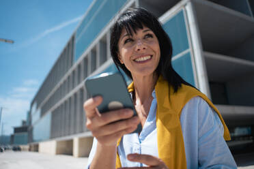 Lächelnde reife Geschäftsfrau mit Smartphone vor einem Bürogebäude - JOSEF19648