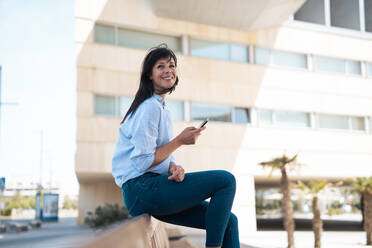 Nachdenkliche Geschäftsfrau mit Smartphone auf einer Bank sitzend - JOSEF19659